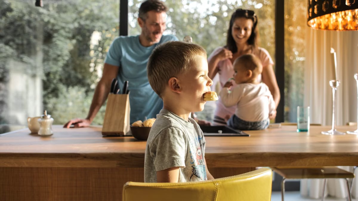 Side view of playful boy eating cookie while sitting at table with parents and sibling on background spending time at home